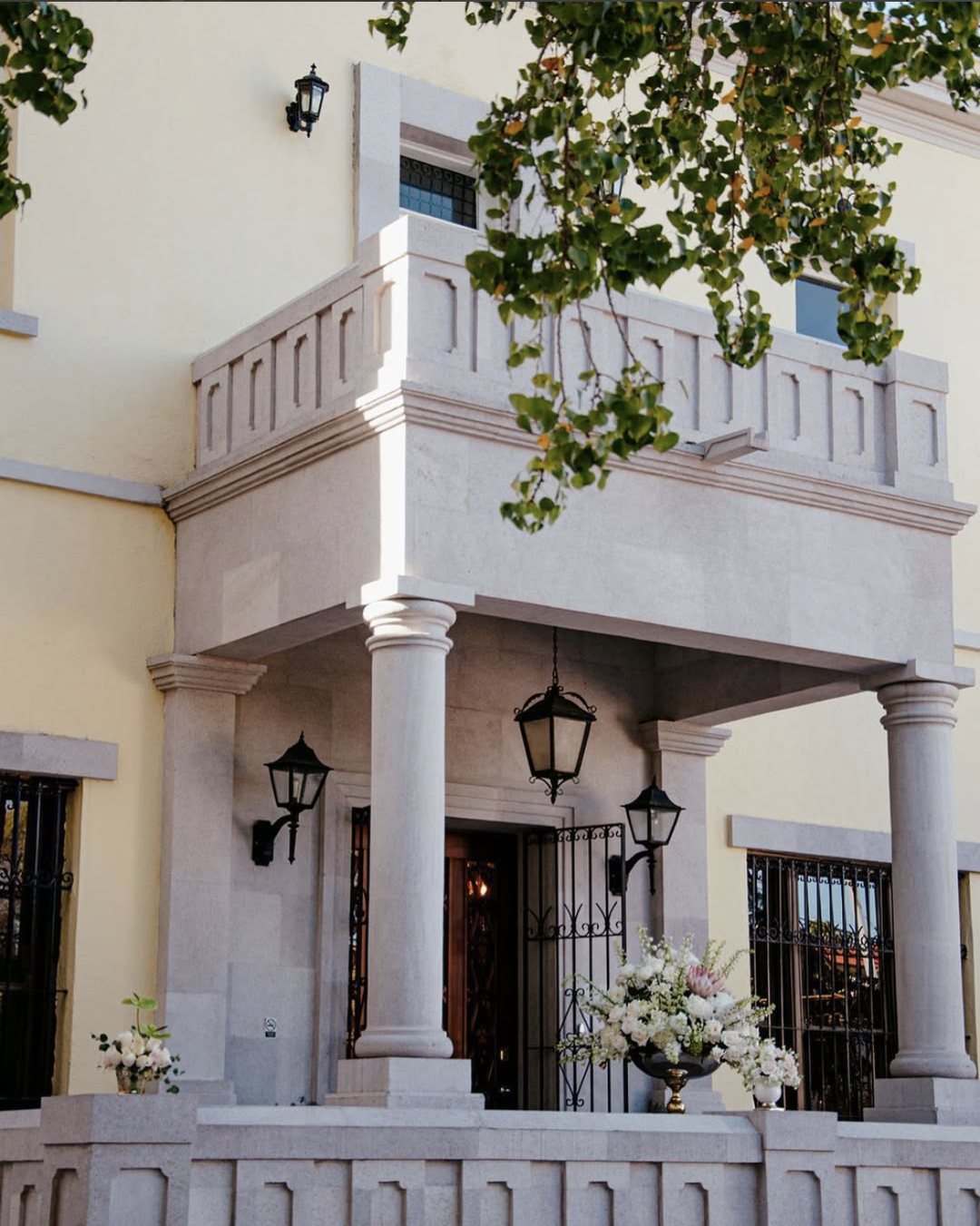 Fachada de Casa Padre Mier: pórtico de cantera con columnas, faroles negros y flores blancas en la entrada